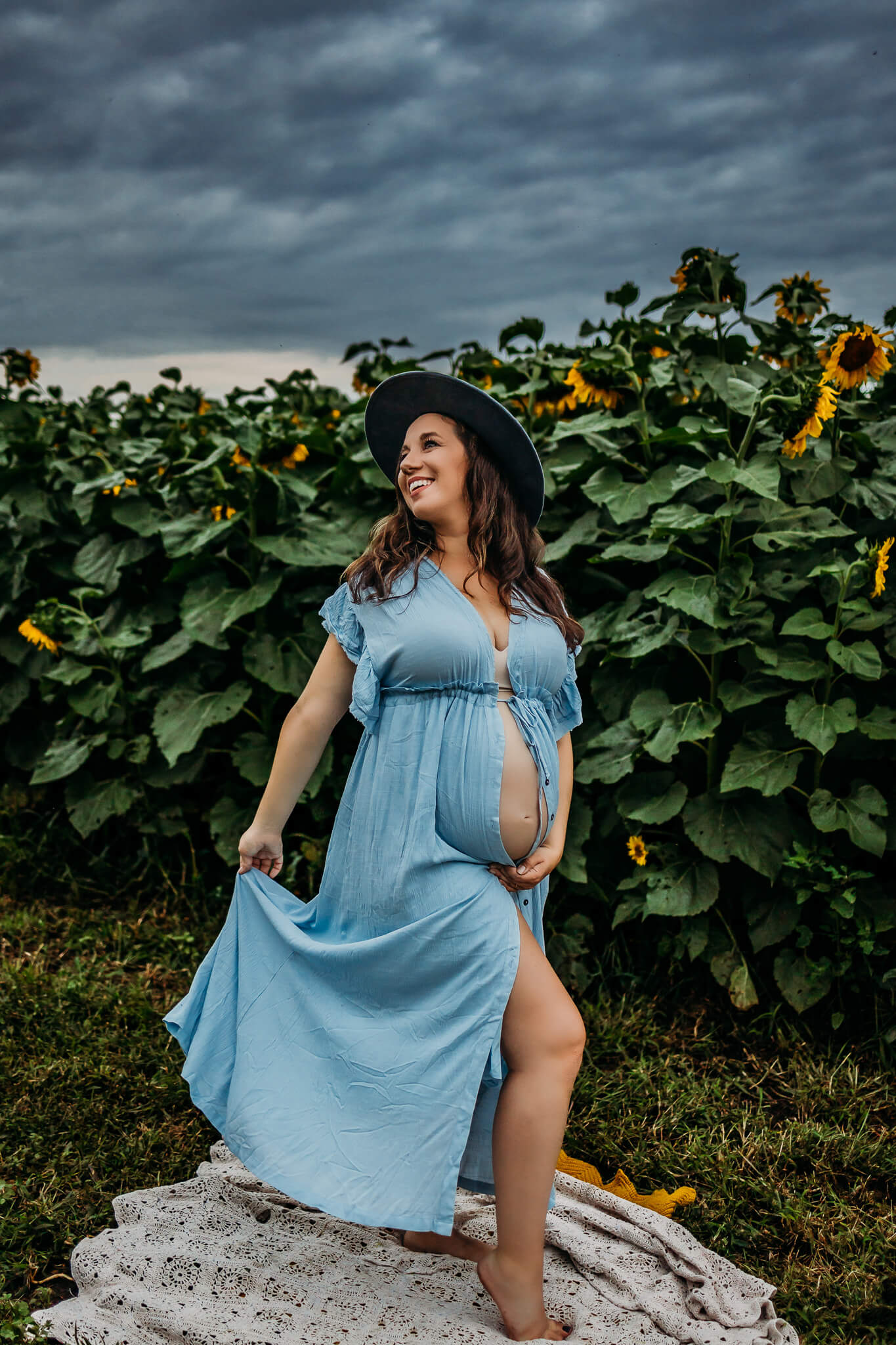 mom to be holding her bump and blue dress while wearing a hat in a sunflower field doula eau claire wi