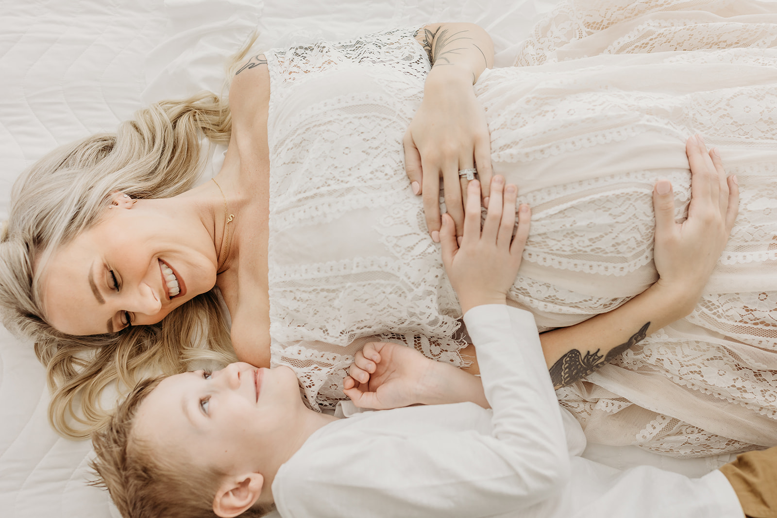 A pregnant mother in a white lace maternity dress lays on a bed cuddling with her older son eau claire chiropractors