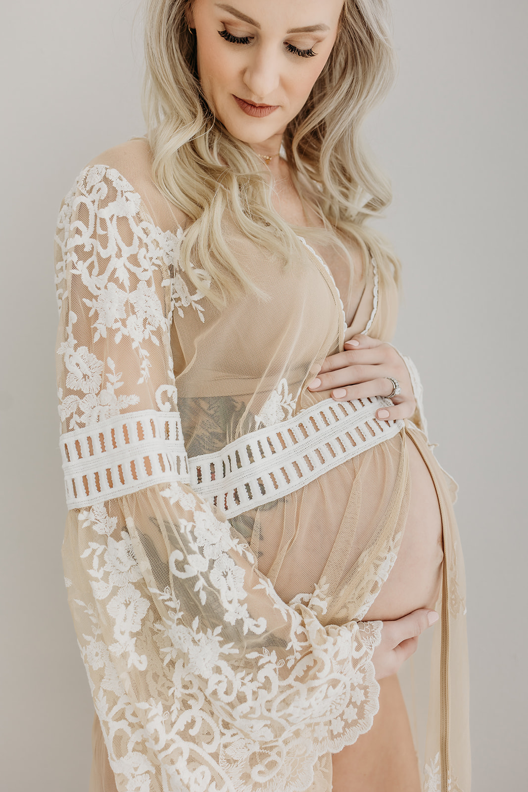 A mother to be in a beige and white lace maternity gown looks down her shoulder while holding her bump in a studio Eau Claire OBGYN