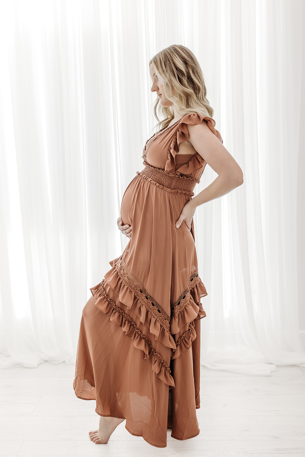 A mother to be stands barefoot in a studio in front of a large window holding her bump in a brown maternity dress HSHS Eau Claire