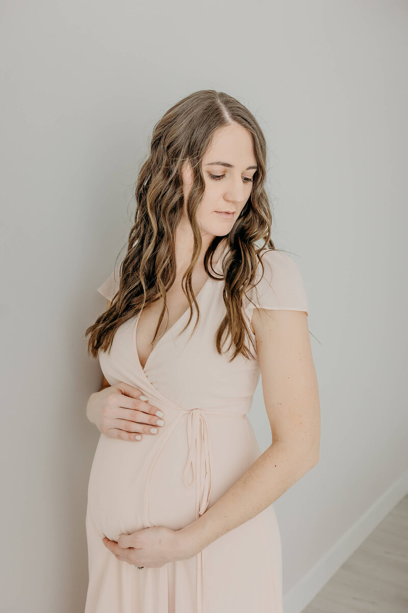 A mother to be stands against a wall wearing a pink maternity gown while looking down her shoulder Marshfield Clinic OBGYN