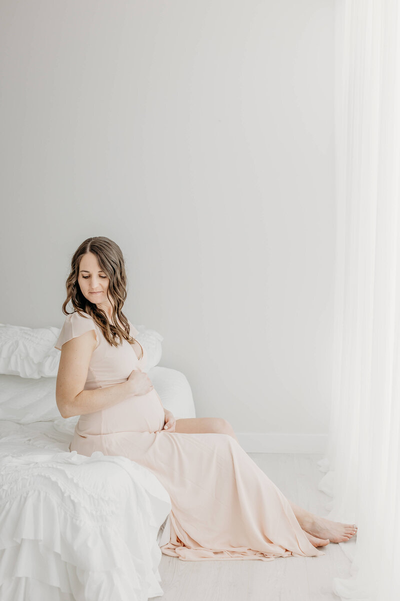 A mom to be sits on the edge of a white bed by a window in a pink maternity dress