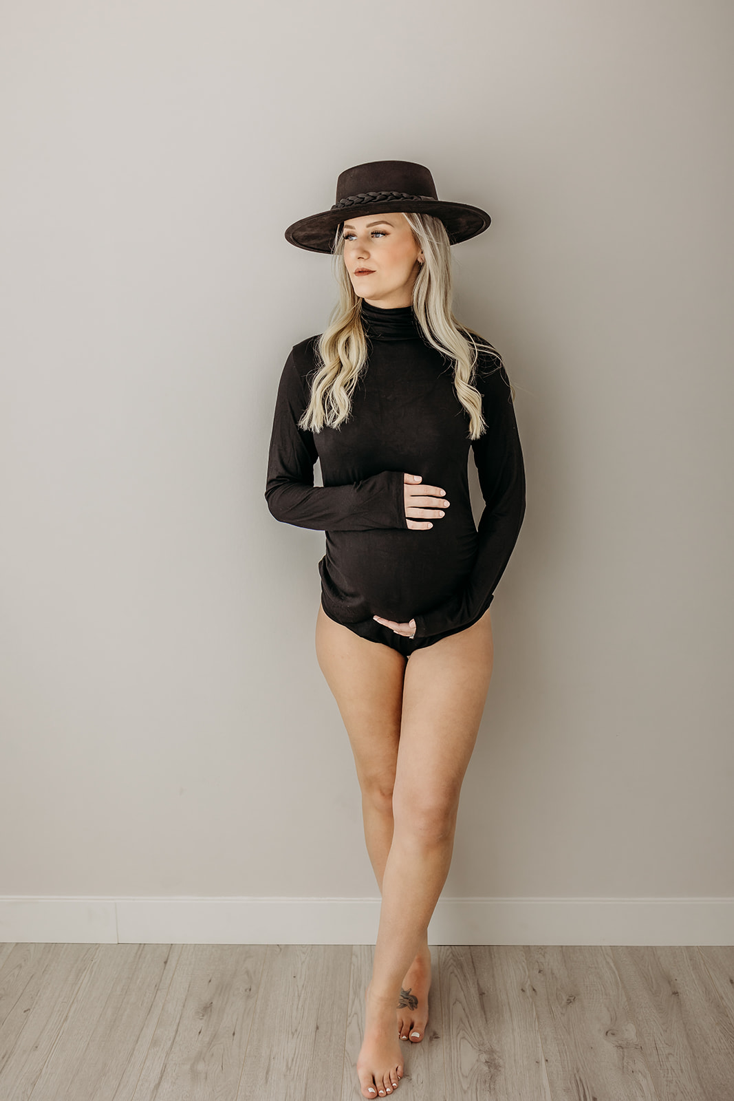 A mother to be in a black long sleeve onesie and hat leans against a wall in a studio Botox Eau Claire