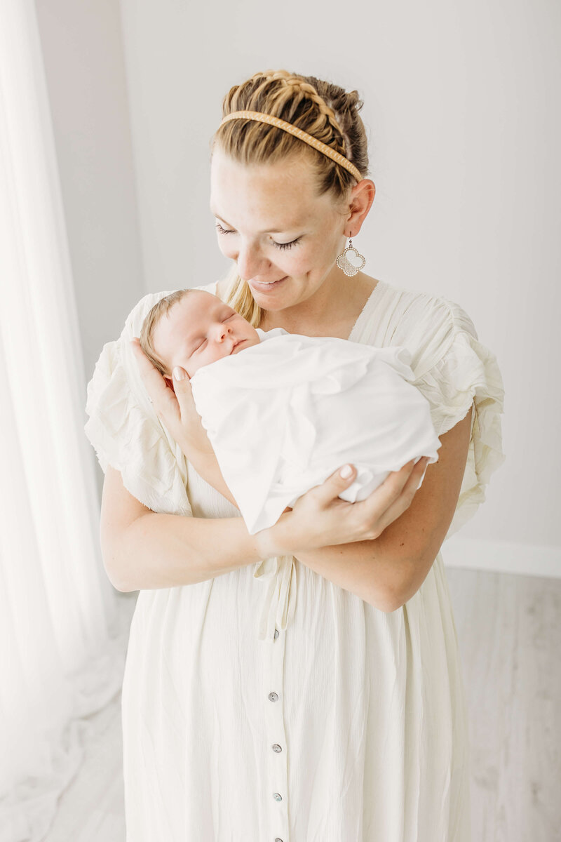 A new mother stands in a studio holding her sleeping newborn baby while wearing a white dress nail salons eau claire