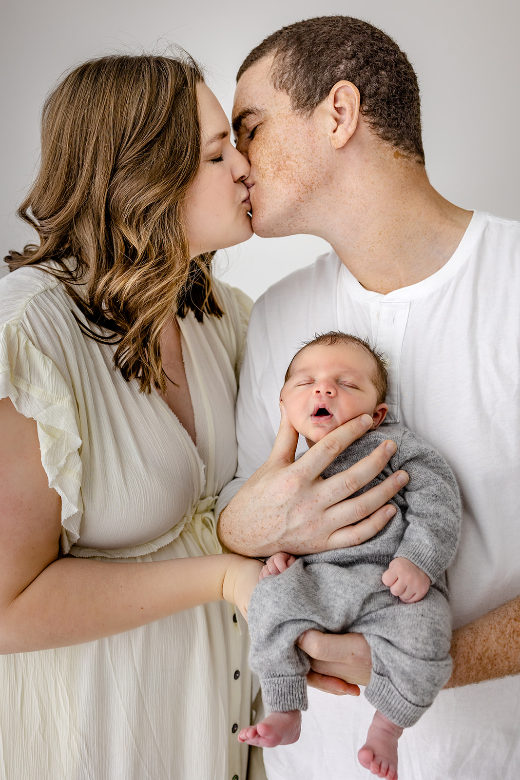 A mom and dad kiss while holding their sleeping newborn baby in a studio before finding a Chippewa Falls Daycare