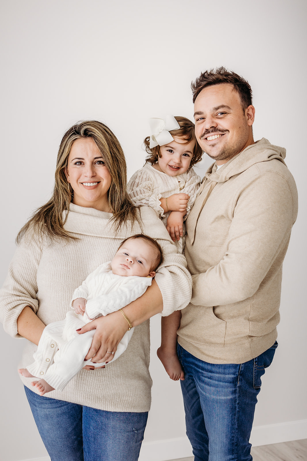 A mom and dad stand in a studio holding their newborn baby and toddler daughter
