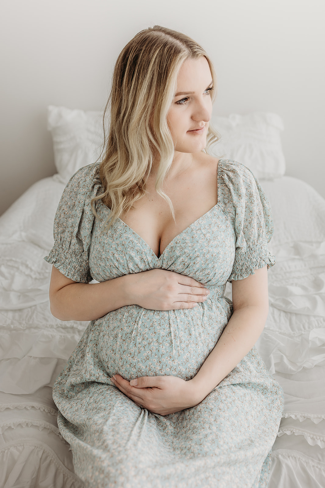 A mom to be sits on a bed in a studio in a green floral dress holding her bump while she looks out a window after visiting Oakleaf Birth Center
