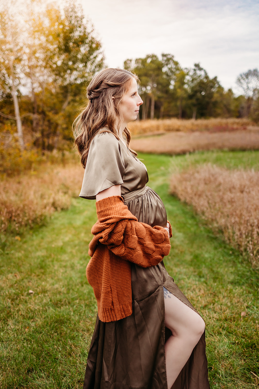 A mom to be in a maternity dress stands in a park trail through tall grass after Prenatal Massage Eau Claire