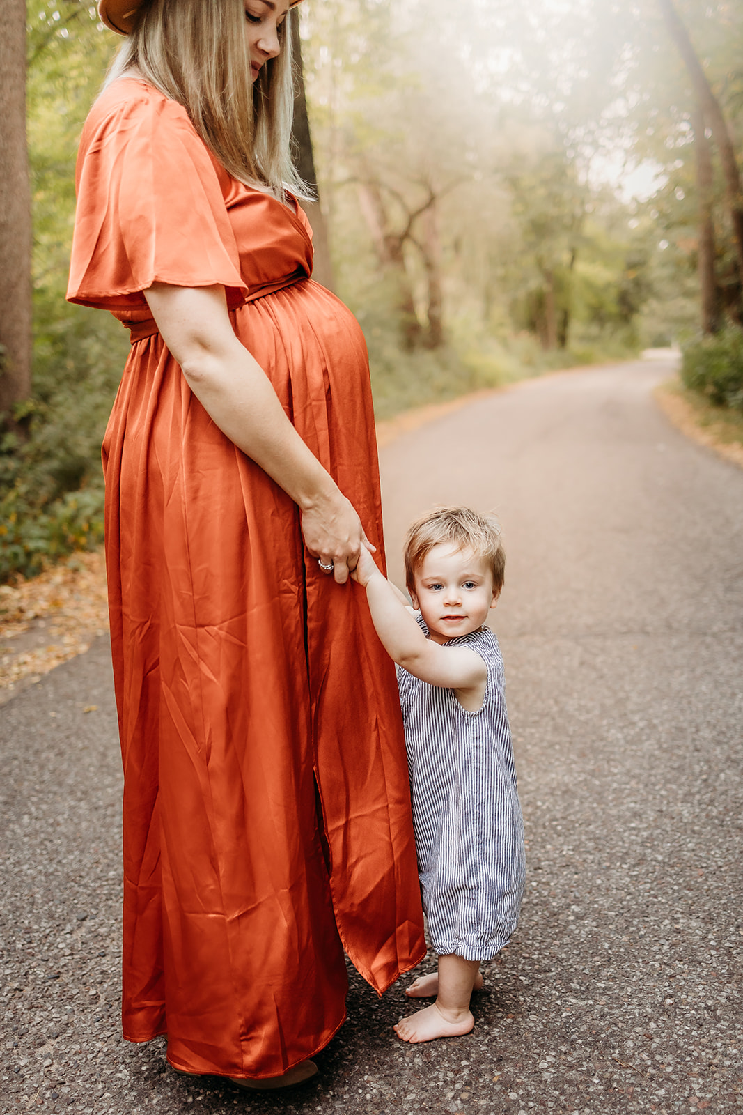 A pregnant mother in a red dress stands with her toddler son in a park sidewalk after Prenatal Yoga Eau Claire