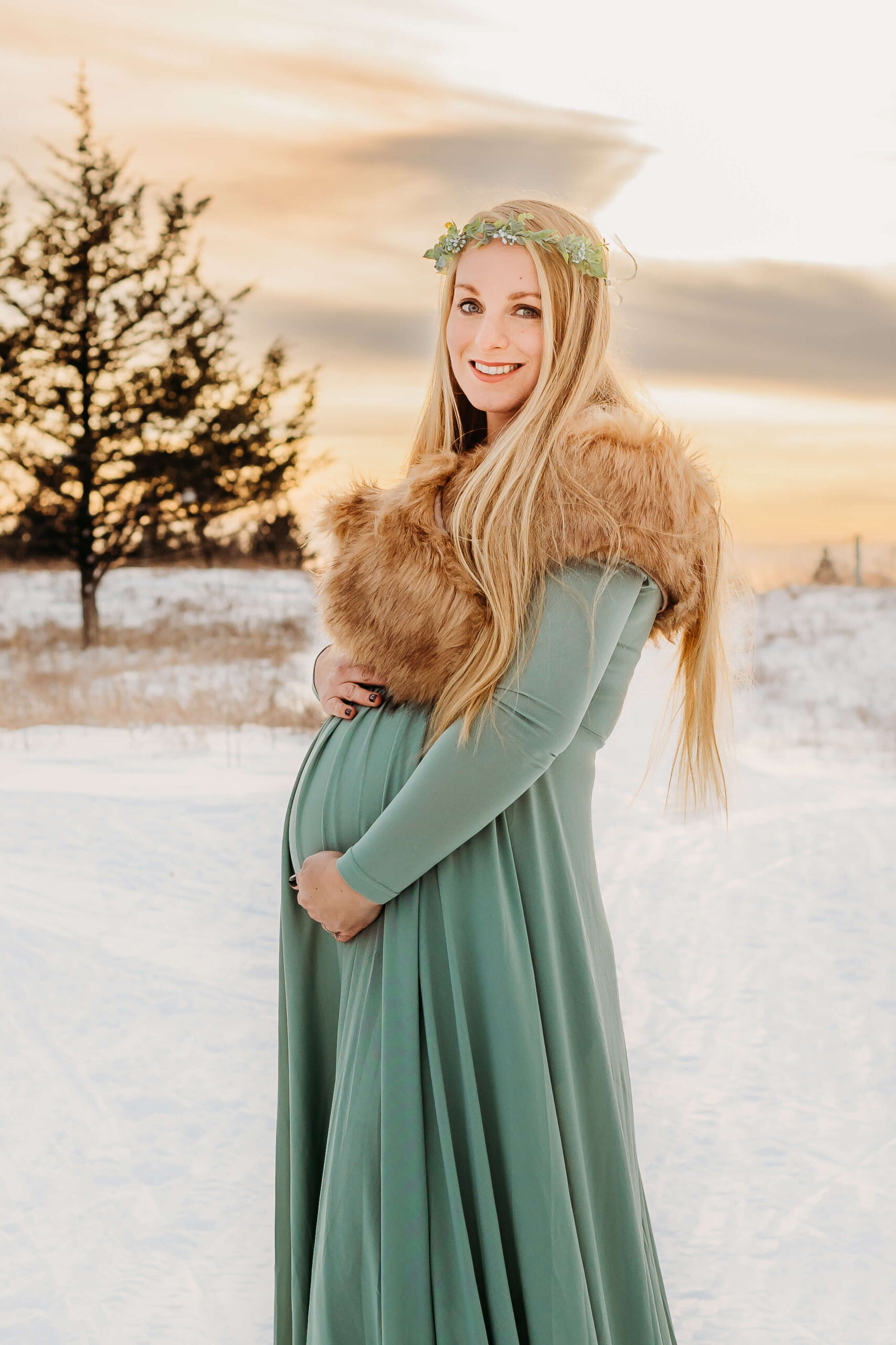 A mother to be holds her bump while standing in a snowy field in a green maternity gown and fur shawl at susnet