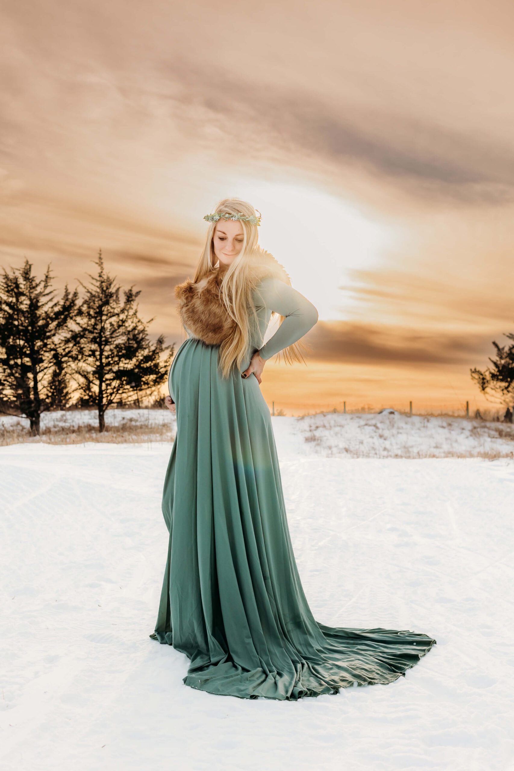 A mother to be rests a hand on her back in a green maternity gown while standing in the snow at sunset after a 3D Ultrasound Eau Claire WI