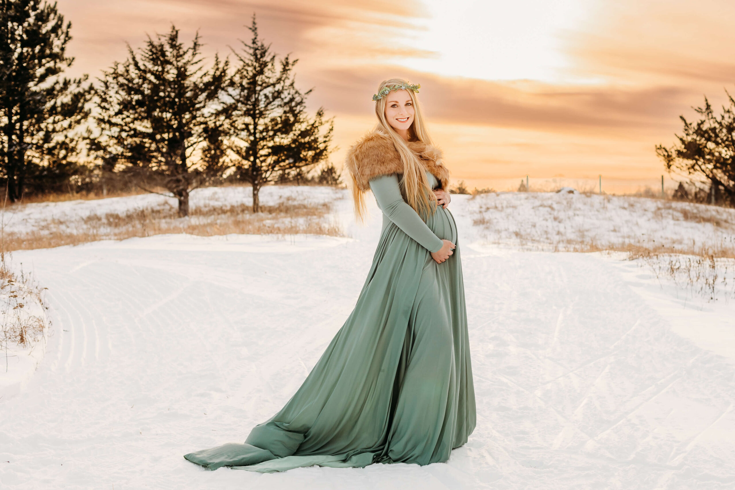 A mother to be in a green maternity gown stands on a snowy hill holding her bump after getting a 3D Ultrasound Eau Claire WI