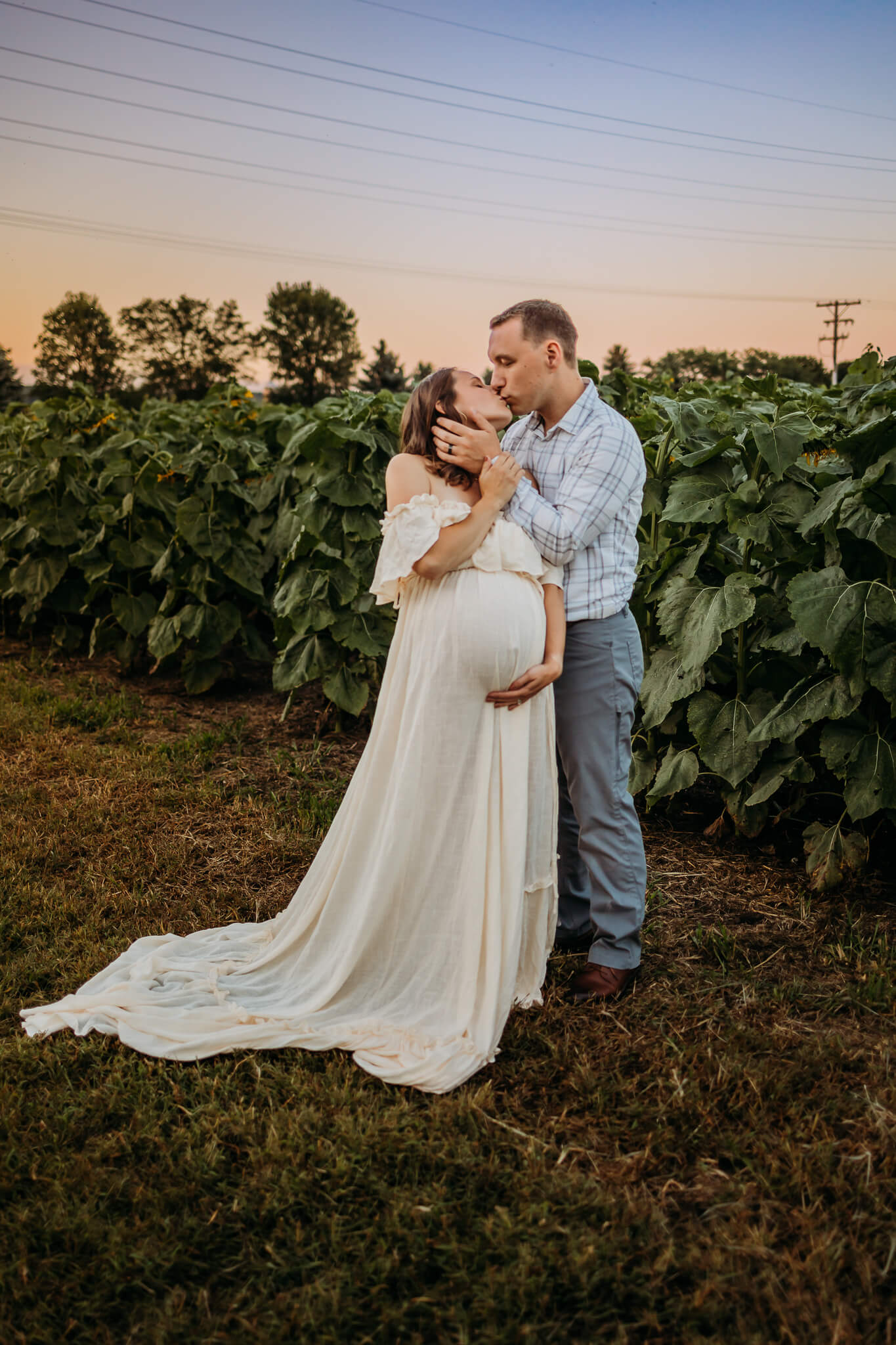 A mother to be in a long white maternity gown leans back for a kiss from her husband in a sunflower field at sunset thanks to acupuncture eau claire