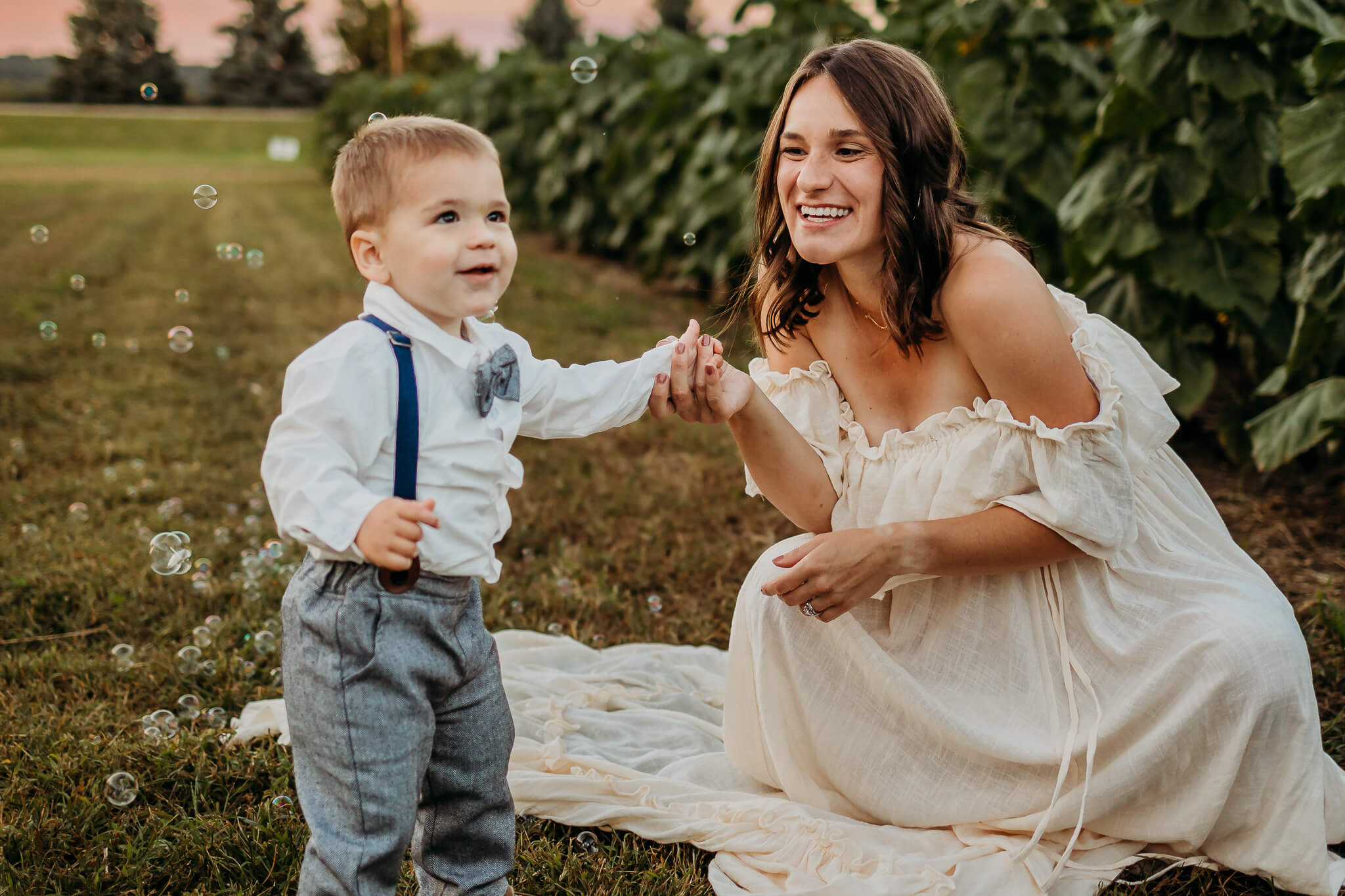 A happy mom plays with bubbles with her toddler son in suspenders and bowtie in a park at sunset thanks to acupuncture eau claire