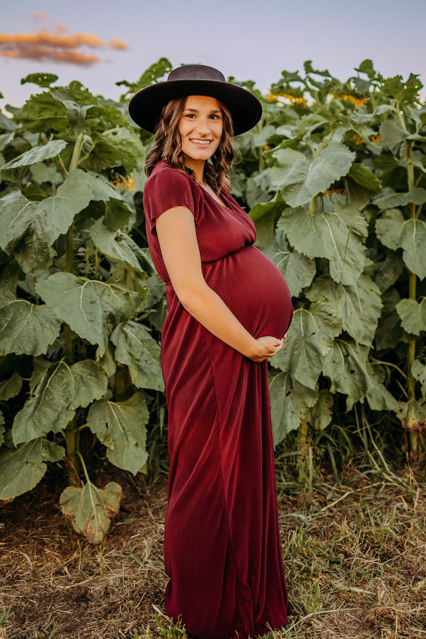 A mom to be wears a red maternity gown in a field of tall sunflowers at sunset