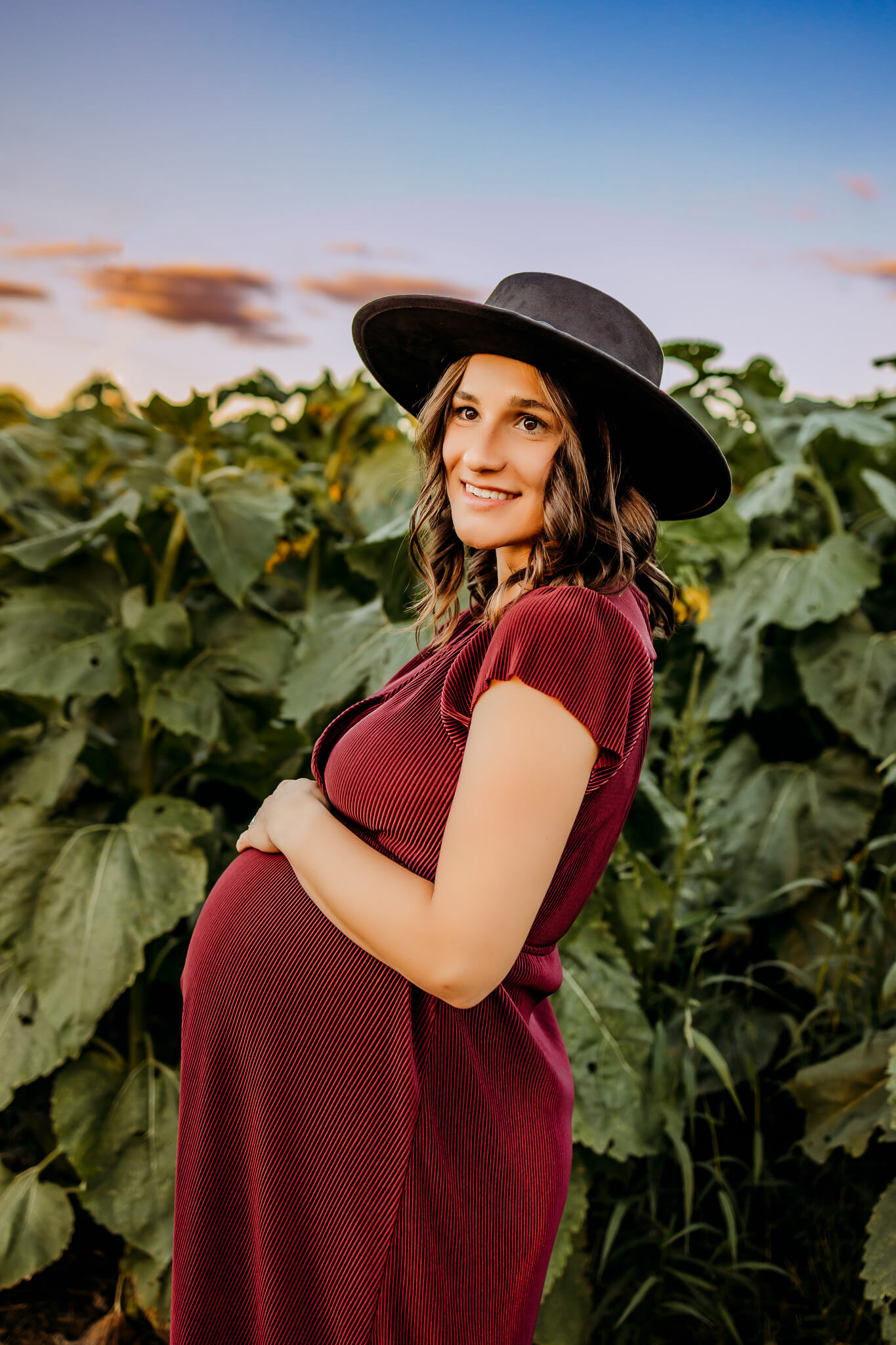 A mom to be in a red maternity dress and large hat stands on a farm at sunset holding her bump after visiting baby shower venues eau claire