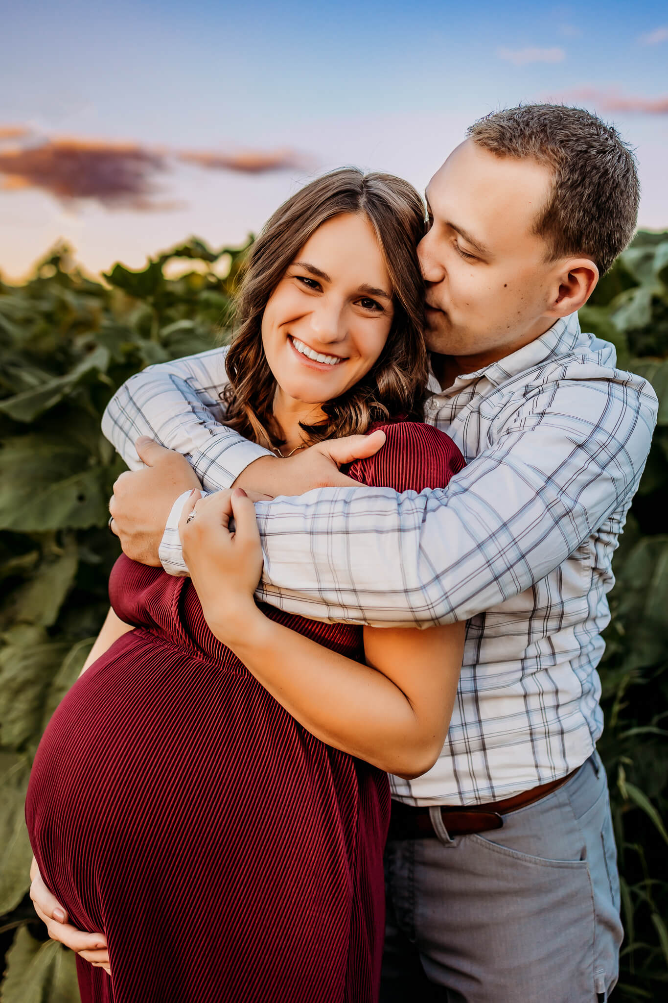 A mother to be in a red maternity dress is hugged and kissed by her husband while standing outside at sunset after visiting baby shower venues eau claire