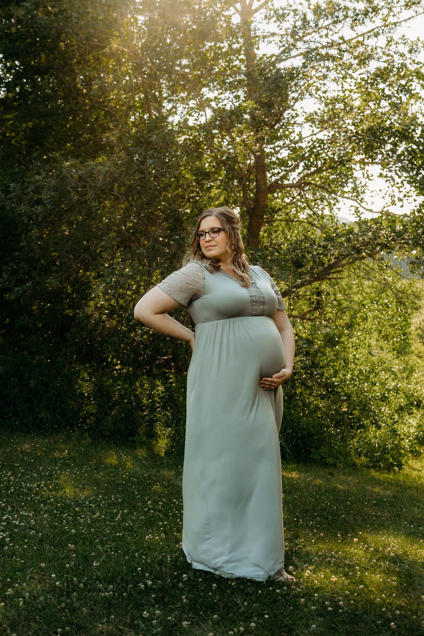 A mother to be in a green maternity gown stands in a forest field at sunset holding her bump