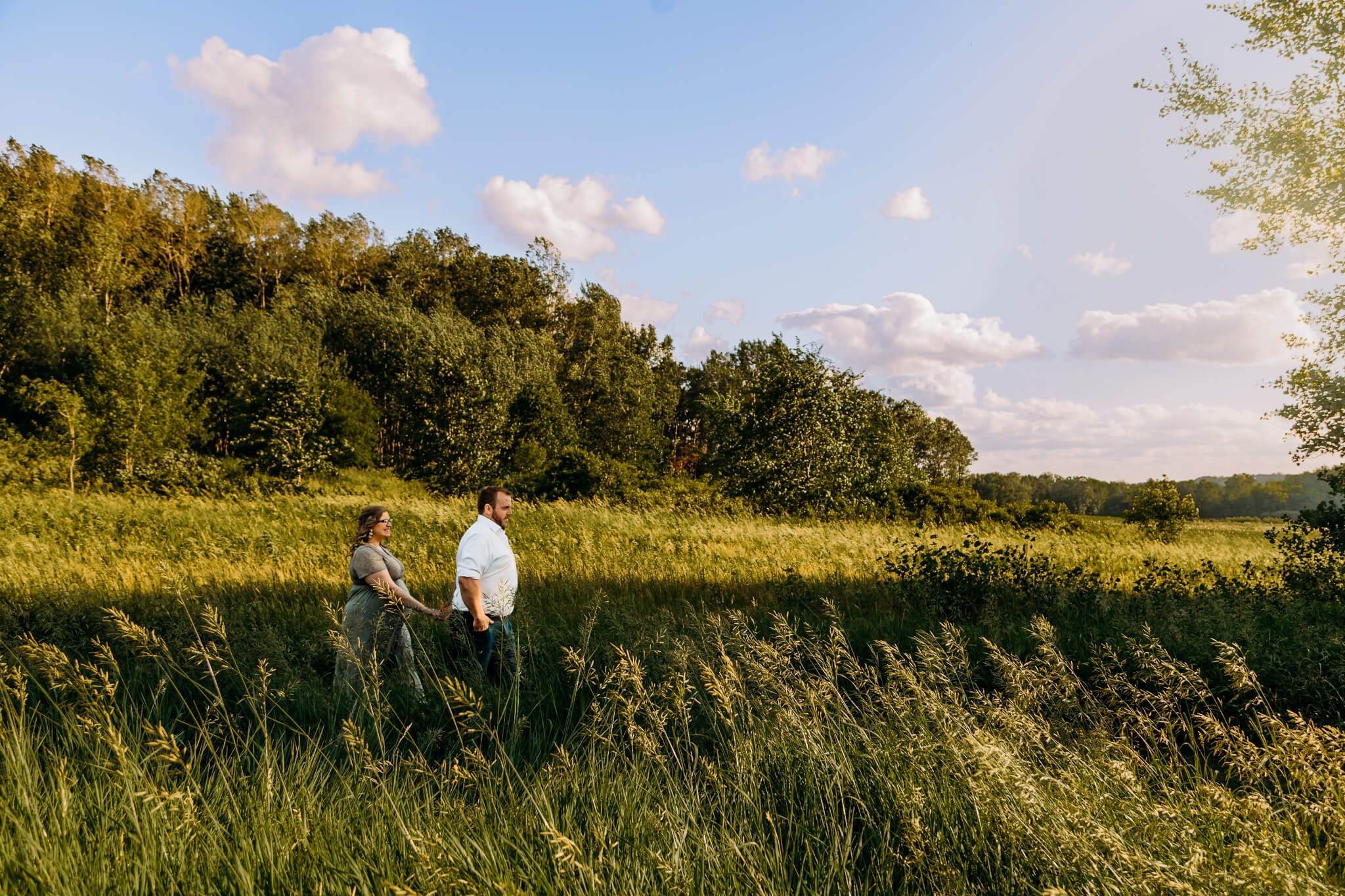A husband leads his pregnant wife through a field of tall grass at sunset after visiting confluence chiropractic