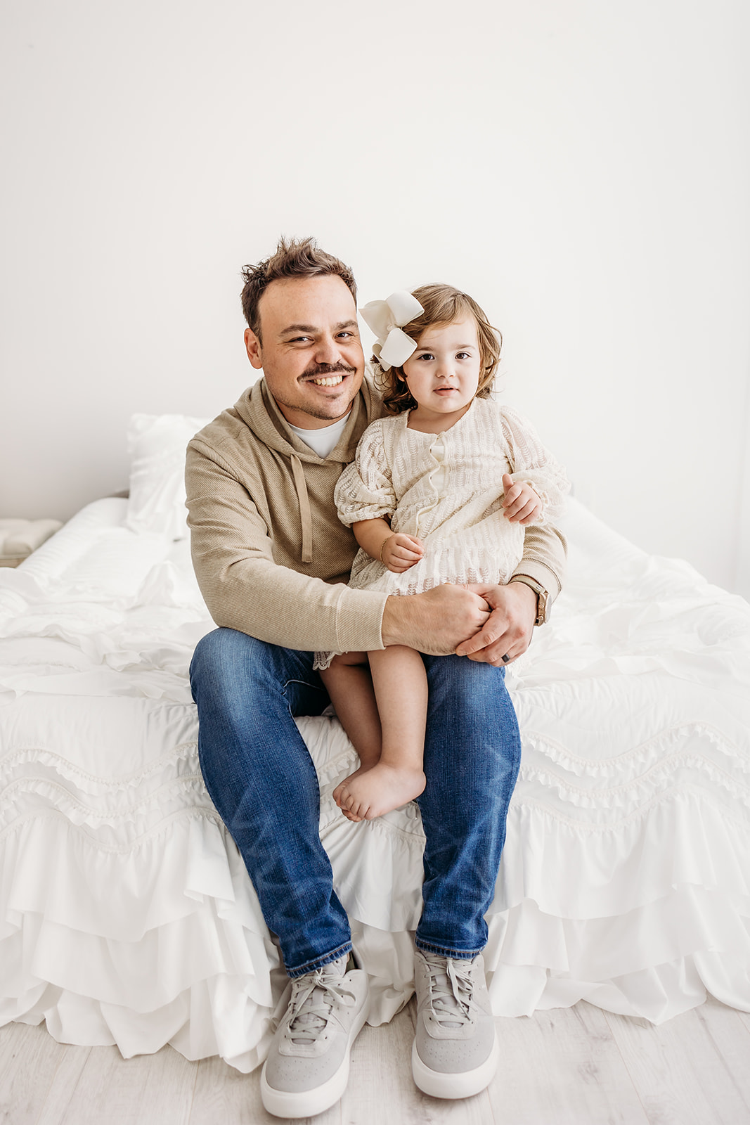 A father in jeans sits on the edge of a bed in a studio with his toddler daughter in his lap