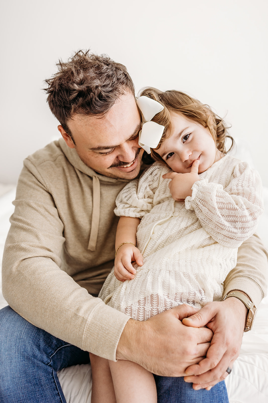 A happy dad sits on a bed hugging his toddler daughter in a studio