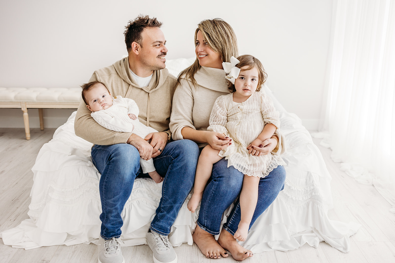 A mom and dad sit on the edge of a bed with their newborn baby and toddler daughter in their laps in a studio before visiting Eau Claire Indoor Playgrounds
