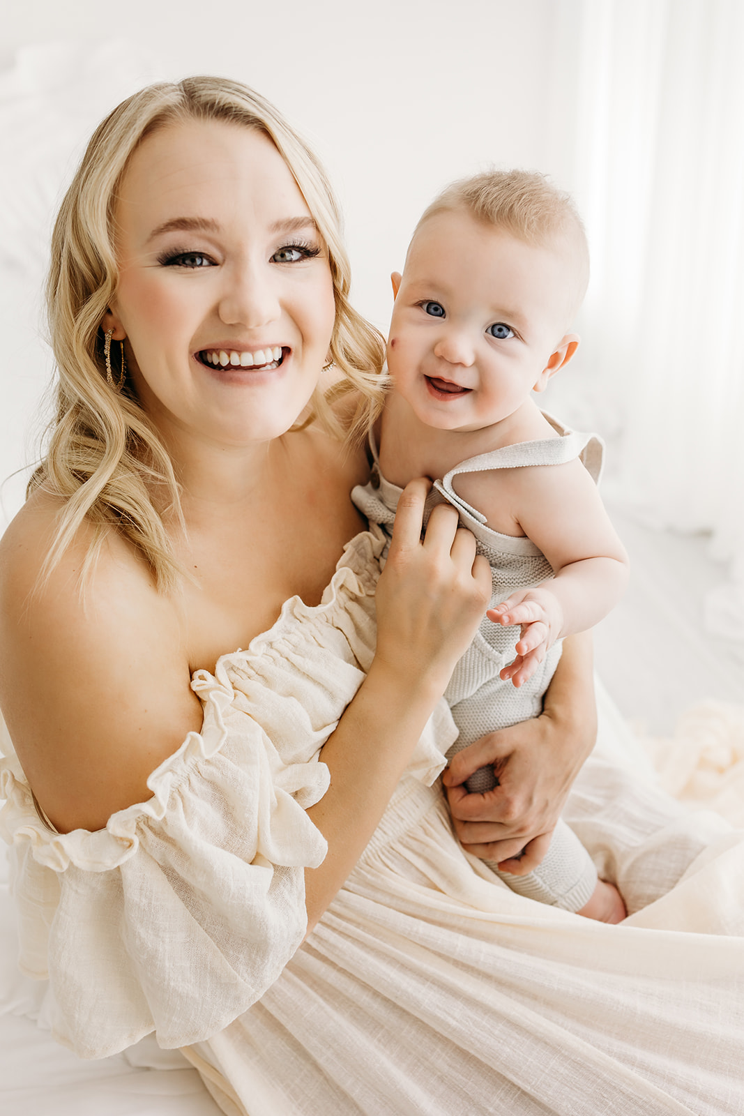 A new mother in a white dress laughs with her infant son in her arms while sitting in a studio