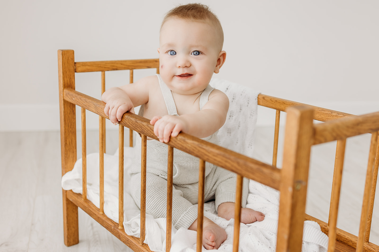 An infant boy in a white knit overalls onesie sits in a wooden crib in a studio after meeting with Mama Bear Lactation Care