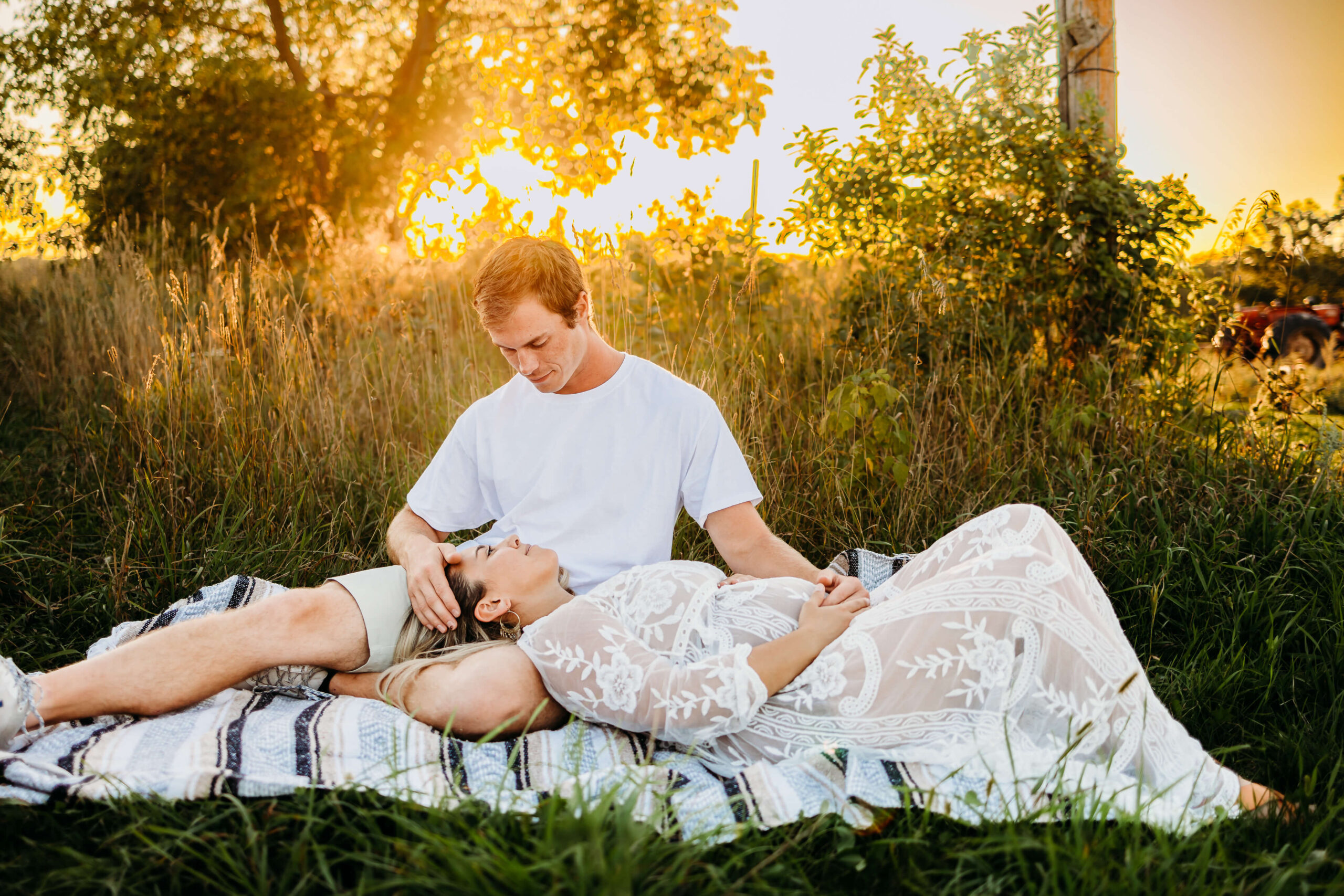 A mom to be in an embroidered maternity dress lays in the lap of her husband on a picnic blanket at sunset
