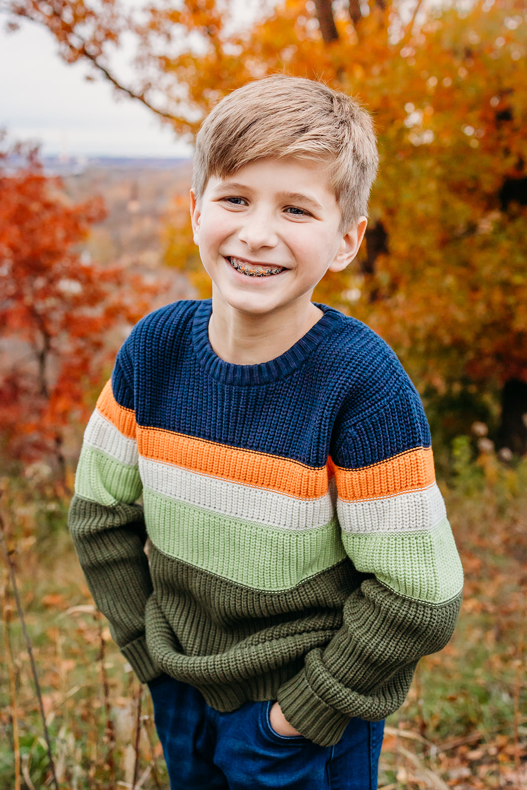 A young boy in a colorful knit sweater stands in a park in fall with hands in his pockets after visiting Pediatric Dentist Chippewa Falls, WI