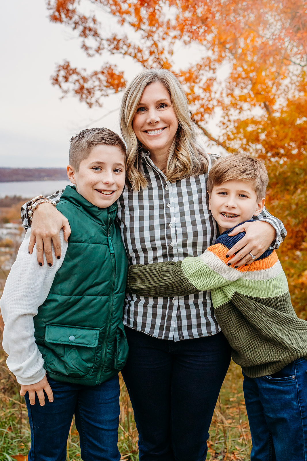 A mother is hugged by her two young sons in a park in fall on a lake before visiting a Toy Store Eau Claire, WI