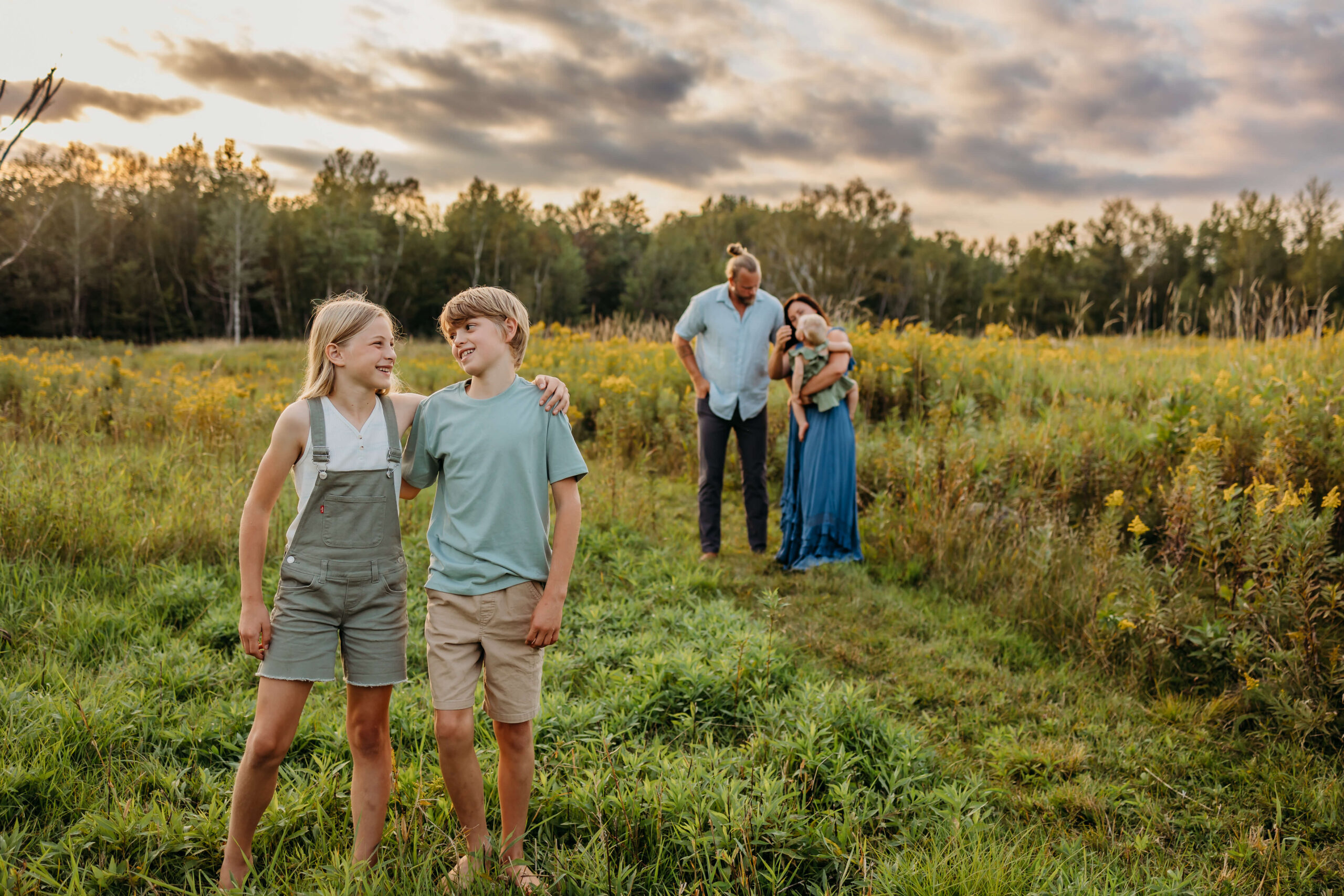 Twin brother and sister stand with arms around each other in a grassy field while mom and dad stand behind them on the trail playing with a toddler before some eau claire summer camps