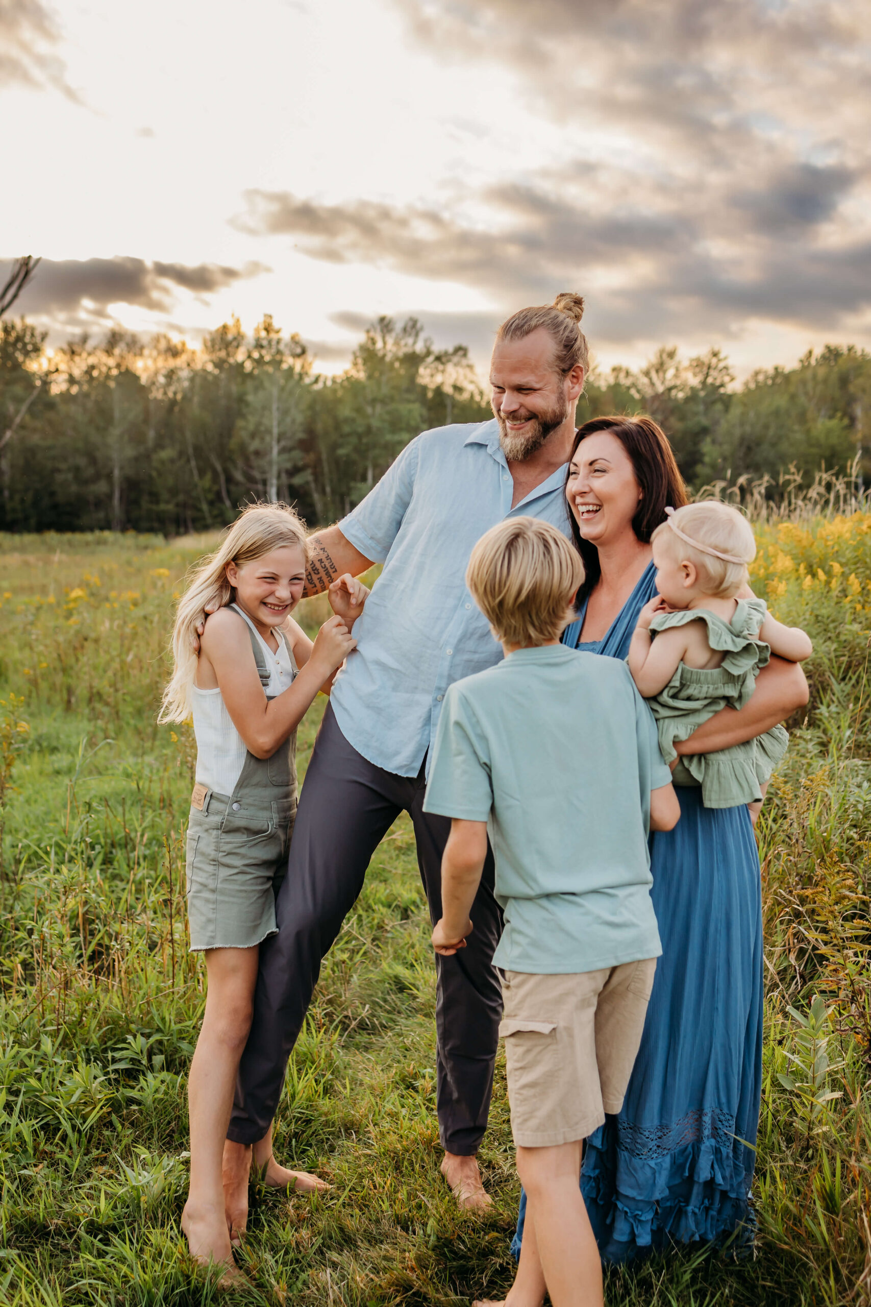 A mom and dad laugh with their two young children and a toddler on mom's hip in a field of wildflowers at sunset before some eau claire summer camps