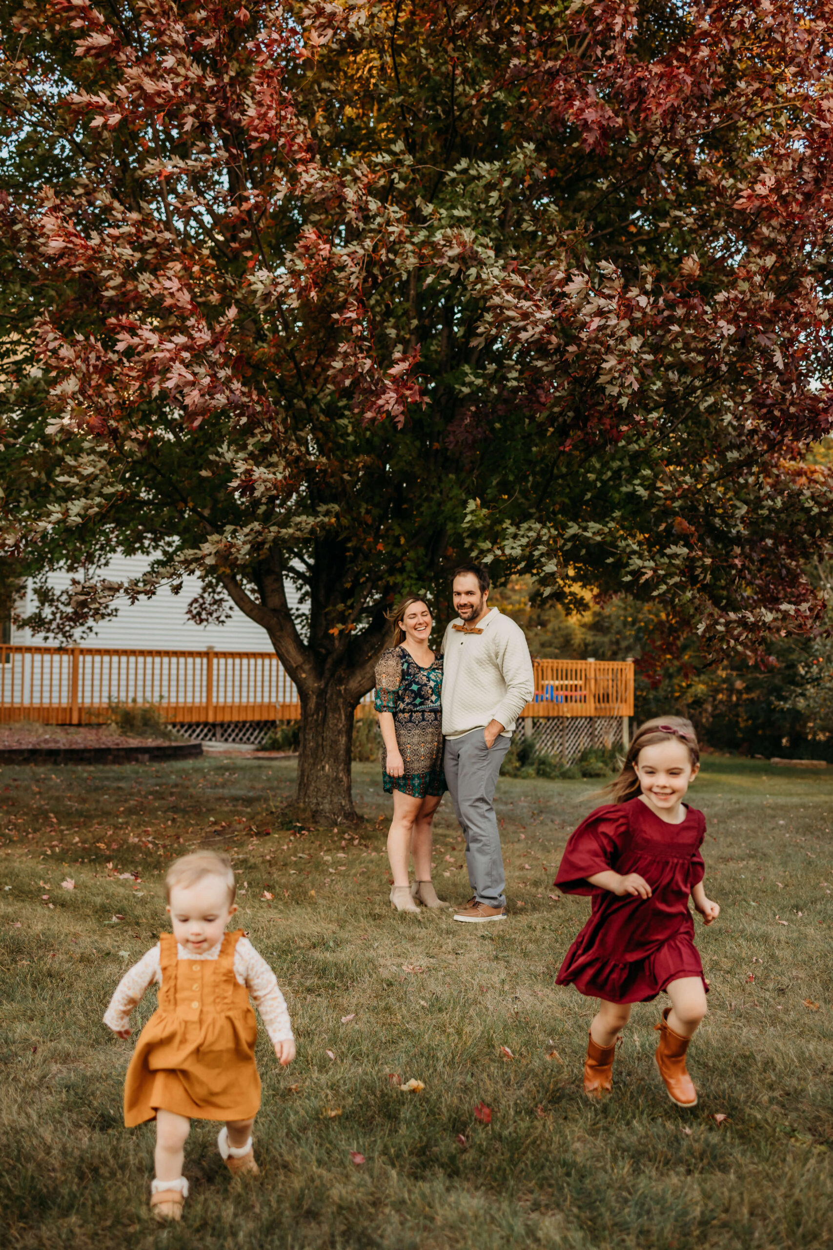 A mom and dad stand under a maple tree as their two toddler daughters in dresses run in front of them before visiting eau claire trampoline park